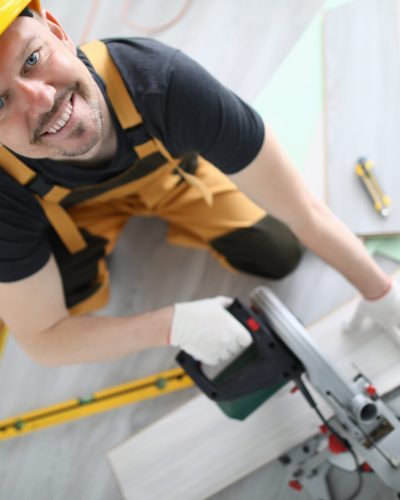Builder in overalls and hard hat smile and look up. Man cut laminate on machine in room.